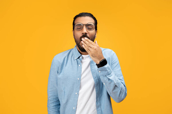 Sleepy tired or bored man in a light blue shirt, covering his mouth while yawning, standing with a tired expression on his face, want sleep against a vibrant yellow background
