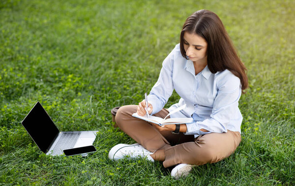 Focused young caucasian businesswoman sitting cross-legged on the grass with a laptop beside her, writing in a notebook, fully absorbed in her work against an urban backdrop