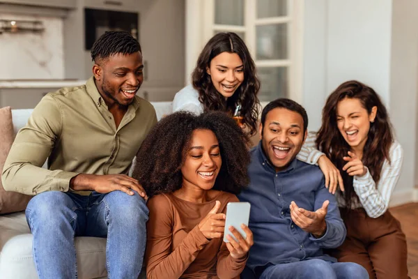 Group of multiethnic friends scrolling through smartphone gathered in modern kitchen, texting and watching funny videos, united in laughter and gadgets leisure during home party