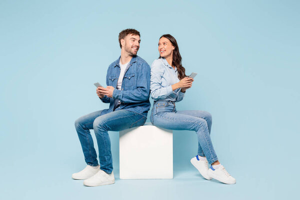 Happy young man and woman sitting back to back, each engaged with their own smartphone, on plain cube against blue background, full length
