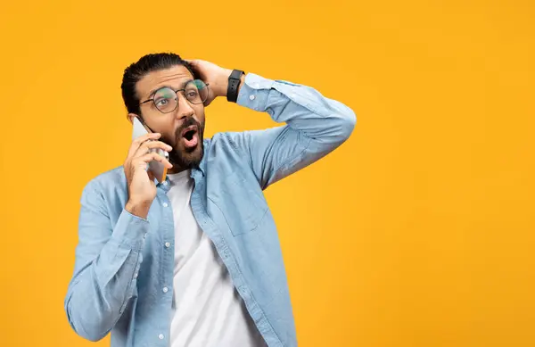 Shocked man in a blue denim shirt talking on a cellphone, holding his head in disbelief or receiving surprising news, with a stunned facial expression against a vibrant yellow background