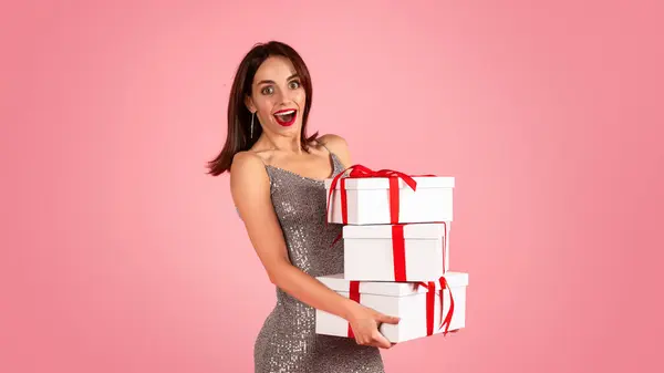 An overjoyed woman with wide eyes and a bright smile, dressed in a glittery silver dress, is carrying a stack of white gift boxes with red ribbons against a soft pink background