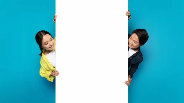 Asian millennial man and woman playfully peeking around a large white vertical banner, with joyful expressions, perfect for advertising space on a bright turquoise background