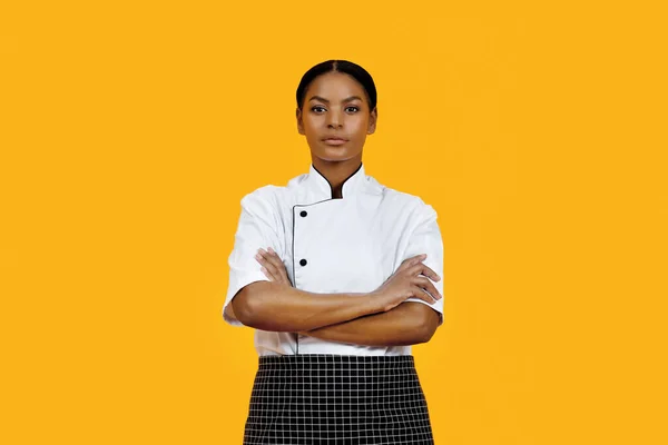 Professional black female chef in white uniform standing with her arms crossed against vibrant yellow background, african american female cook exuding confidence and expertise, copy space