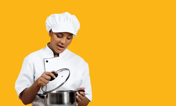 Excited black female chef carefully looking into stainless steel pot, professional african american cook woman in uniform anticipating culinary results, posing isolated on yellow studio background