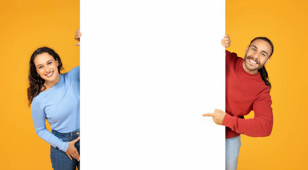 Happy millennial couple cheerful european man and woman showing white blank advertising board between them, pointing at placard with mockup copy space, orange background