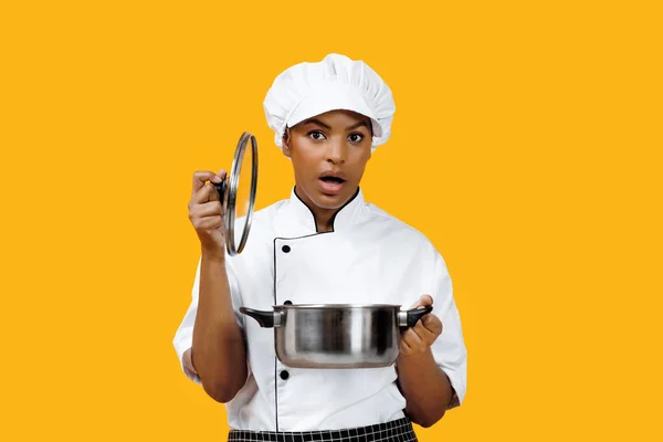 Surprised black female chef lifting the lid off pot and looking at camera, african american cook woman capturing her unexpected reaction to the dish, standing against bright yellow background