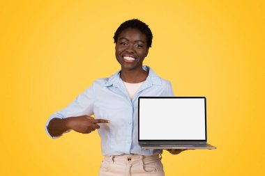 Glad young african american woman student in casual point finger at computer laptop with empty screen, isolated on yellow studio background. Study and education app, attention website for lesson