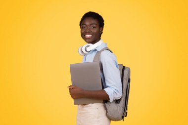 Smiling teen african american lady student in wireless headphones, hold laptop, ready to lesson, isolated on yellow studio background. Study and education with device at school