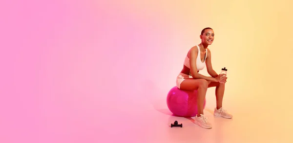 A smiling woman with a shaved head takes a break on a fitness ball, holding a water bottle, wearing a white sports bra and shorts, in a gym setting with a pink and yellow background