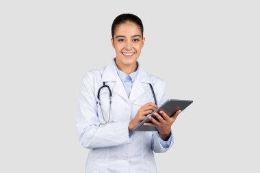 Steteskoplu neşeli Avrupalı bir bayan doktor, geleneksel tıp ve modern teknoloji ile sağlık hizmetlerinin kesişimini somutlaştıran bir dijital tablet tutuyor.
