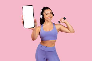 Dambılı ve kulaklıklı neşeli, formda bir bayan pembe arka planda duran, boş ekranlı, kocaman bir telefon gösteriyor. Spor ve spor için tavsiye başvurusu