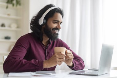 Bir fincan kahve ve kulaklıklı neşeli Hintli adam iyi aydınlatılmış ev ofisinde dizüstü bilgisayarıyla çalışırken gülümsüyor. Doğulu serbest çalışan mutlu bir erkek.