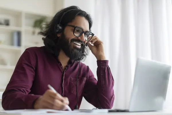 Online Education. Indian Smiling Man In Headset Study With Laptop At Home, Handsome Young Eastern Guy Sitting At Desk, Looking At Computer Screen And Taking Notes, Enjoying Distance Learning
