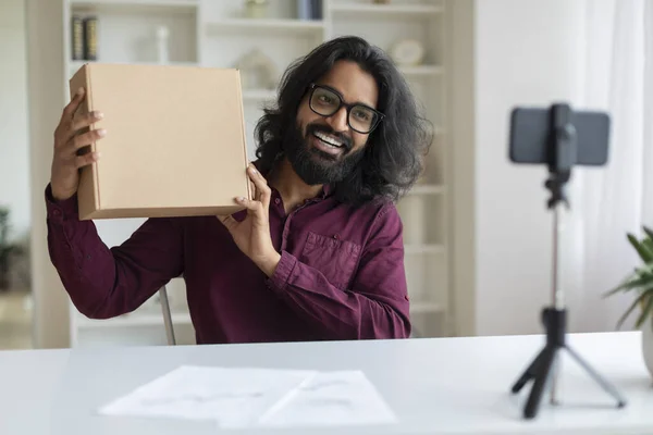 Happy Indian man presenting cardboard box to smartphone camera, recording unboxing review video while sitting at desk at home, smiling eastern male using mobile phone on tripod for capturing content