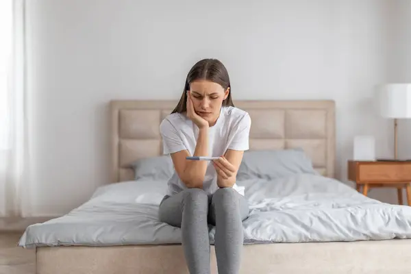 令人失望的女士举行怀孕测试 一个人坐在床上卧室的内部 女性健康问题 意外怀孕和分手后的孤独 — 图库照片