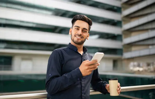 Successful arab businessman in black shirt typing on smartphone near modern office building, standing with takeaway coffee and smiling to camera with confidence. Application for business purpose