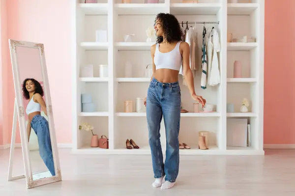 Chic young latin woman in white crop top and high-waisted jeans looking at her reflection in full-length mirror in room with soft pink tones