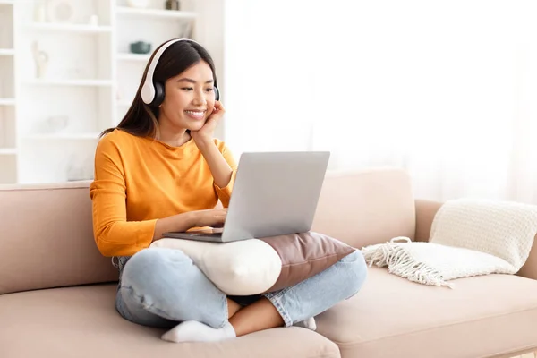 Relaxed young asian woman watching movie on laptop, sitting on couch in cozy living room, enjoying favorite podcast, playing video game, using wireless headphones, copy space