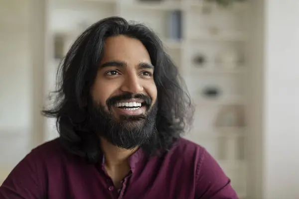 Portrait of handsome young indian male looking aside and smiling, closeup shot of happy eastern man daydreaming in home interior, bearded millennial guy embodying spirit of happiness and contentment
