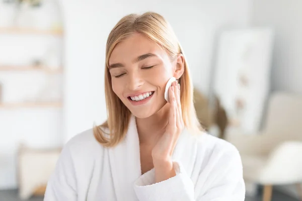 Smiling millennial woman with blonde hair moisturizing her face with a toner using cotton pad, embodying pure refreshment and natural beauty in modern bathroom interior. Beauty and cosmetics