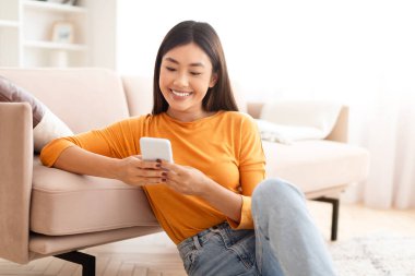 Hoş, genç Asyalı bir kadın evinde koltuğun yanında oturuyor, telefon kullanıyor ve gülümsüyor, arkadaşlarına veya sevgililerine mesaj atıyor, en yeni mobil uygulamalardan hoşlanıyor, kayıyor, internetten alışveriş yapıyor, kopyalama boşluğunu kopyalıyor.