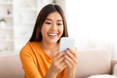 Gülümseyen Asyalı genç bir kadının evde akıllı telefon kullanması, sosyal ağlara mesaj atması ya da göz gezdirmesi, kanepede dinlenmesi, kopyalaması. Mobil uygulama, cihaz bağımlılığı