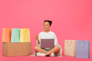 Çevrimiçi alışveriş için dizüstü bilgisayar ve kredi kartı kullanan Mutlu Asyalı Kadın, Pembe Stüdyo Arkaplanındaki Parlak Alışveriş Torbaları Arasında oturan Koreli Alışverişkolik Kadın, İnternet Alışverişlerinin keyfini çıkarıyor