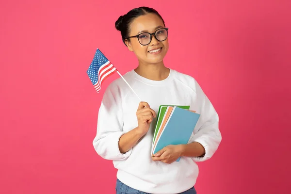 笑着的亚洲女学生自豪地举着美国国旗和彩色笔记本 快乐的年轻韩国女学生代表着国际教育和文化交流 站在粉色的背景上 — 图库照片