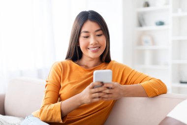 Evdeki modern akıllı telefonu kullanan, sosyal medyada veya internetten alışveriş yapan, rahat oturma odasındaki kanepede oturan Asyalı genç ve güzel bir bayanın samimi fotoğrafı.