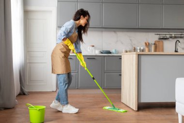 Zemini temizlemek için yeşil paspas kullanan bej önlüklü ve sarı eldivenli konsantre genç kadın, ahşap döşemeye de aynı kovayı koyuyor.