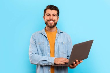 Dizüstü bilgisayarıyla poz veren neşeli adam mavi stüdyo arka planında kameraya gülümsüyor. Pozitif serbest çalışan genç adam defteri online iş için hazır tutuyor, portre çekimi için.