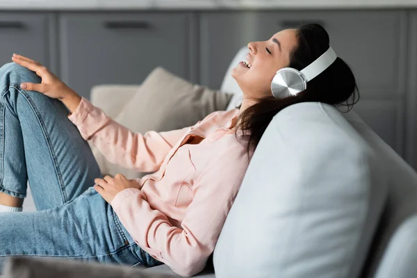 Blissful woman reclines on sofa, losing herself in music with stylish white headphones, her expression one of pure joy and relaxation in her serene home