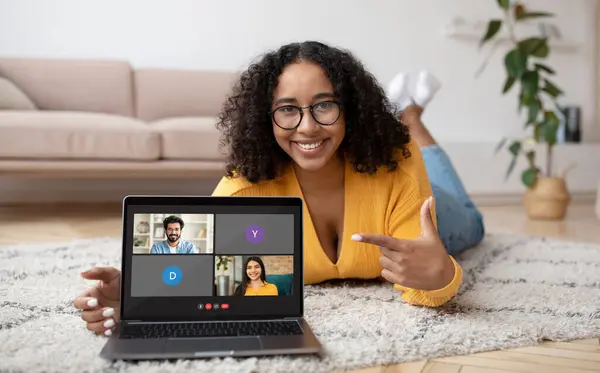 Telecommunication concept. Cheerful millennial black woman wearing eyeglasses laying on floor at home, show laptop computer with video call screen, have online meeting with friends