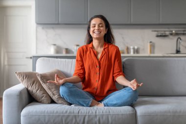 Huzurlu genç kadın, Lotus pozisyonunda meditasyon yaparken rahat gri koltukta huzur buluyor, modern yaşam alanındaki sükunet ve rahatlığı somutlaştırıyor.