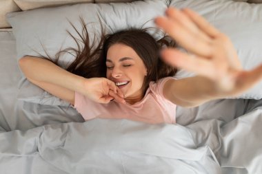 Joyful young lady in pajams stretching in bed with closed eyes and smile on her face after waking up in the morning at home, top view clipart