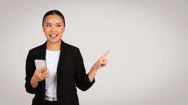 Siyah ceketli, kendinden emin bir iş kadını sağ elini gösterirken sol elinde bir akıllı telefon tutuyor ve tarafsız bir arkaplana gülümsüyor. İş ve iş