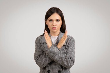 Houndstooth blazer ceketli kararlı genç bir kadın hiçbir olumsuz hareket yapmaz, düz bir arka planda ciddi bir ifadeyle ellerini çapraz yapar