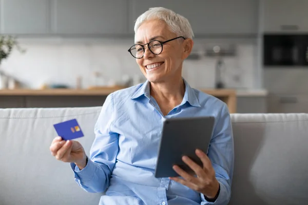 Online Shopping. Cheerful mature woman holding debit credit card and websurfing on digital tablet, ordering purchases and clothes via internet, sitting on sofa in living room interior