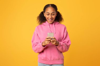 Sarı stüdyo arkaplanının üzerinde duran akıllı telefondan sosyal medyaya kayan siyahi genç kız. Pembe kapüşonlu genç bayanın cep telefonuyla mesajlaşması, harika haberler okuması.