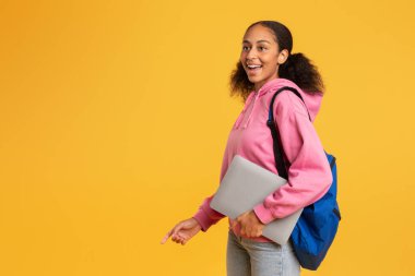Heyecanlı Afrikalı Amerikalı öğrenci dizüstü bilgisayarını ve sırt çantasını sarı stüdyo arka planında taşıyor ve öğrenim döneminin başlangıcından duyduğu mutluluğu dile getiriyor. Harika bir eğitim teklifi. Boşluğu kopyala