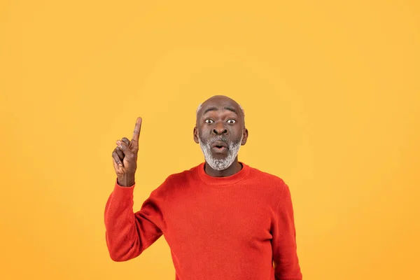Surprised and excited glad elderly man in a red sweater pointing upwards, with a wide-eyed expression of amazement against a vivid yellow background, studio, free space