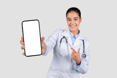 Gülümseyen genç Avrupalı doktor boş ekranlı bir cep telefonu gösteriyor, sağlık uygulamaları veya tıbbi danışma hizmetleri için mükemmel, gri stüdyo arka planında izole edilmiş