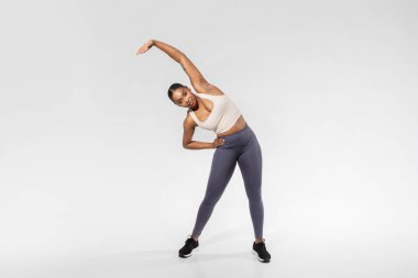 Spor Giysili Genç Siyah Kadın Kolunu Kaldırırken Çekirdeği Beyazların Arkaplanına Yana Eğ, Stüdyo Çekimi. Afrika kökenli Amerikalı kadın antrenmandan önce ısınma egzersizi yapıyor. Boşluğu Kopyala