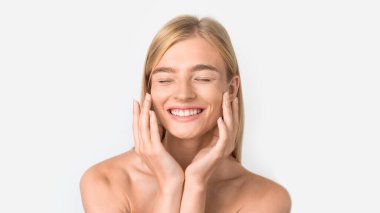 Genç, beyaz tenli, cildine özen gösteren, kozmetik ve nemlendirici kullanan, gözleri kapalı, yanaklarına dokunan ve beyaz arka planda gülümseyen sarışın kadın. Cilt bakımı ve hijyen. Panorama