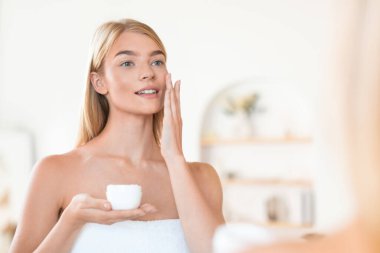 Çekici sarışın bayan cildine nemlendirici sürerken evde banyo yaparken cildine bakım yapıyor ve sabahları havluya sarılı olarak güzelliğini şımartıyor.