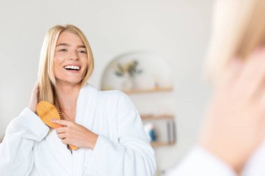 Neşeli sarışın milenyum hanımefendisi aynaya bakıp gülümserken saç bakım ayininin tadını çıkarıyor. Ev banyosunda güçlü uzun saçlarını tarıyor. Kendini pohpohlama ve güzellik. Boş alan