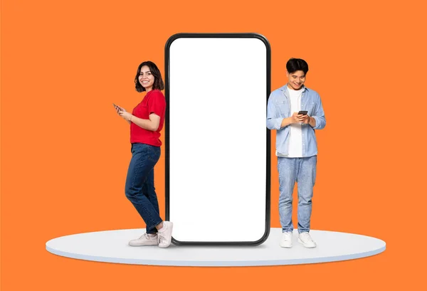 スマートフォンにカジュアルに焦点を当てた若い男女 アプリやインターフェイスモックアップに最適な巨大な空白のスマートフォン画面の横に立って 活気に満ちたオレンジ色の背景に設定 — ストック写真