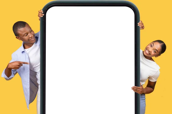 Excited black man and woman holding gigantic smartphone frame, pointing to the blank screen with enthusiasm, ideal for app or advertisement display on yellow background