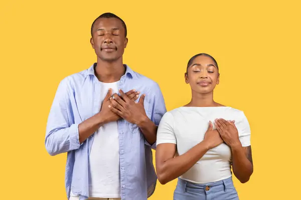 Peaceful black couple stands with hands over their hearts and eyes closed in gesture of gratitude and reflection, set against calming yellow background